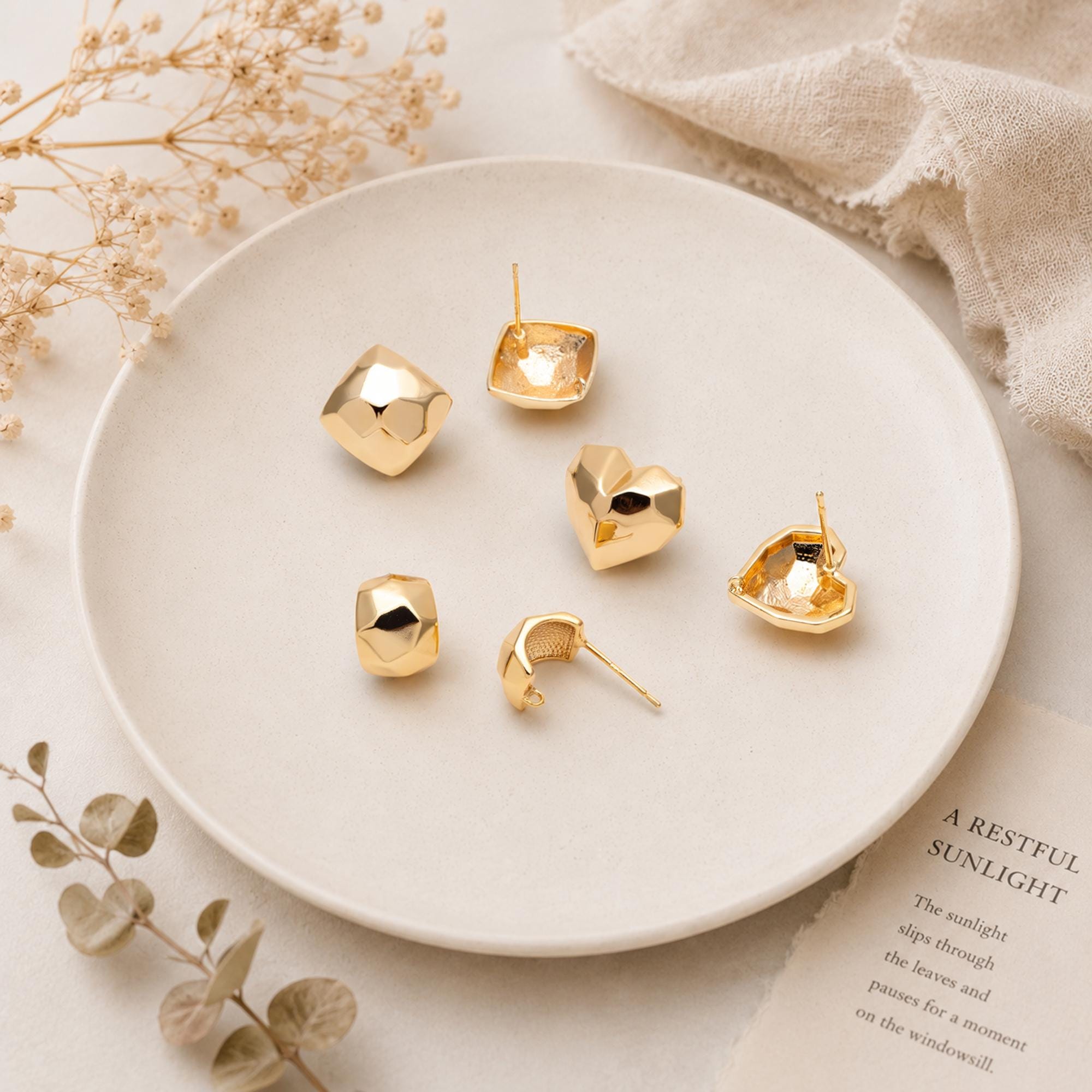May include: A collection of gold-colored earrings in various geometric shapes, including squares, hearts, and rounded forms. The earrings are displayed on a white plate, with a soft, neutral background. The text "A RESTFUL SUNLIGHT" is visible in the background.